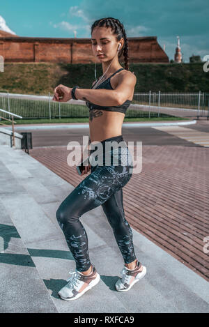 Atletica Giovane bambina guarda l'orologio, misure di tempo di impulso, fitness, sportswear, gambali e top. A portata di mano, telefono ascolta musica cuffie. In Foto Stock