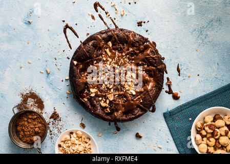 Torta al cioccolato con le nocciole su sfondo blu con il cacao in un colino e nocciole tritate in una ciotola Foto Stock