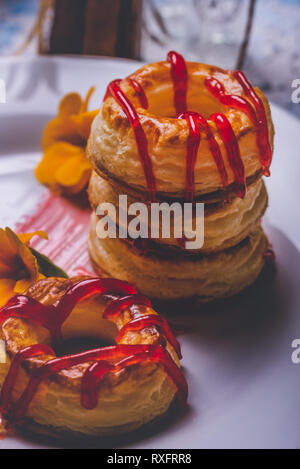 Foto verticale con varie ciambelle preparate dalla pasta sfoglia di pasta alimentare. Rosso fragola topping è versato sul ring. Le ciambelle sono poste su piastra bianca con pochi Foto Stock