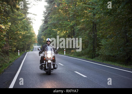 Barbuto biker in occhiali da sole, casco e pelle nera di equitazione abbigliamento moto cruiser lungo la strada asfaltata avvolgimento tra alti alberi verdi sul soleggiato su Foto Stock