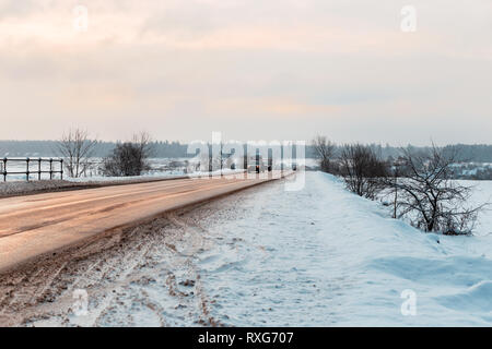 Inverno strada tra i campi in campagna. La riflessione del sole in asfalto, sabbia sulle banchine e spazi aperti nella neve Foto Stock