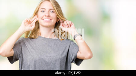 Giovane e bella donna che indossa sovradimensionare t-shirt casual su sfondo isolato sorridente tirando le orecchie con le dita, funny gesto. Problema di Audition Foto Stock