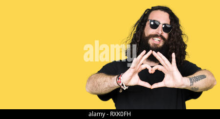 Tanga giovane uomo con i capelli lunghi e la barba indossando occhiali da sole sorridente in amore che mostra il simbolo del cuore e la forma con le mani. Concetto romantico. Foto Stock