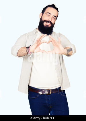 Giovane uomo con i capelli lunghi e la barba e orecchini sorridente in amore che mostra il simbolo del cuore e la forma con le mani. Concetto romantico. Foto Stock