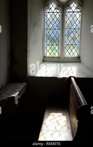 Luce che risplende attraverso una finestra di vetro colorato su una vecchia chiesa pew e pietra ogham paleocristiano di pietra scolpita St Odoceus Chiesa Llandawke Galles Foto Stock