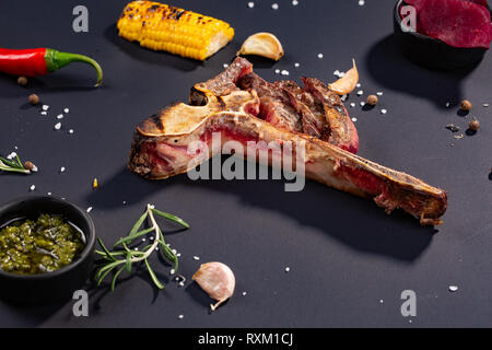 Set di grigliate a fette T-bone steak con grigliate di mais, salsa, peperoncino, rosmarino, sale, aglio su sfondo nero. Graduale aggiunta di pezzi di carne Foto Stock