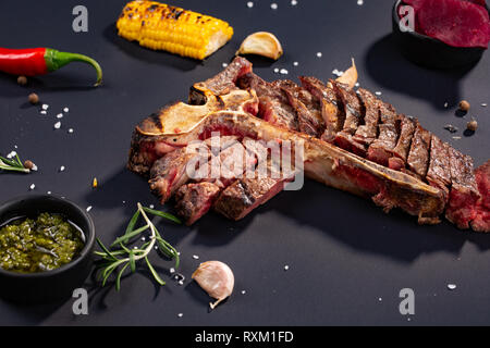 Set di grigliate a fette T-bone steak con grigliate di mais, salsa, peperoncino, rosmarino, sale, aglio su sfondo nero. Graduale aggiunta di pezzi di carne Foto Stock