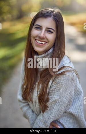 Attraente di moda giovane donna nella luce della sera retroilluminati da sole lateralmente in piedi guardando la telecamera con un bel sorriso amichevole in un autu Foto Stock