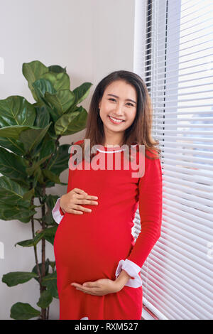 Bella donna incinta in abito rosso in posa a decorate studio, sorridente alla fotocamera. La mamma in piedi vicino alla finestra. Foto Stock