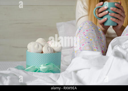 Mani femminili in possesso di una tazza con il drink nel letto. Marshmallows e meringhe in una scatola. Buona mattina e concetto di rilassamento. Spazio di copia Foto Stock