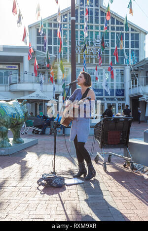 Giovane e bella bruna ragazza o donna che gioca la sua chitarra e canto come un suonatore ambulante di strada o un artista interprete o esecutore al V&A Waterfront, Città del Capo, Sud Africa Foto Stock