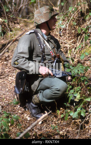 Un WW2 soldato tedesco indossa periodo WW2 uniforme egli si inginocchia nel bosco tenendo un Sten mitragliatrice. Foto Stock