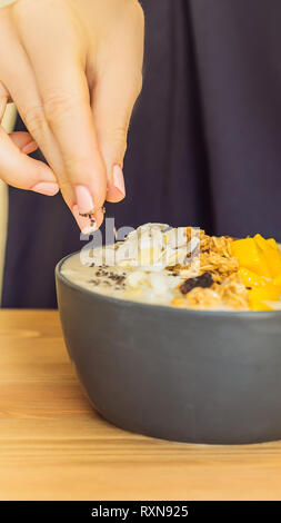 Donna frullato di cuochi bocce fatta con mango, banana, granola, cocco grattugiato, dragon frutta, semi di Chia e menta su sfondo di legno. Concetto di frutta Foto Stock