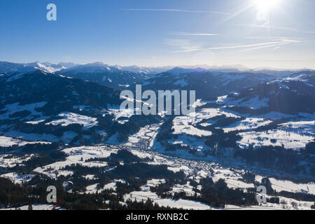 Vista da un drone su un inverno montagne coperte di foreste e di neve nelle Alpi e piccoli villaggi in distanza. Foto Stock