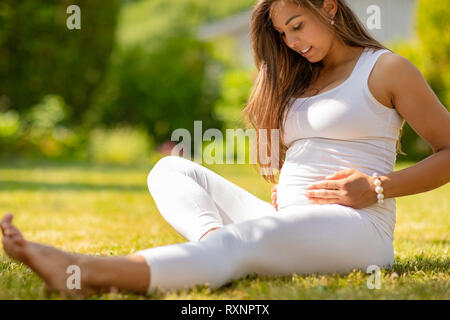 Premurosa donna incinta seduta sull'erba nel suo giardino Foto Stock
