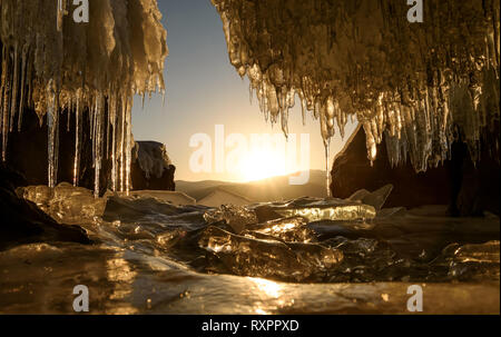 Incredibile sunrise oltre le montagne e il lago ghiacciato in inverno dalla caverna di ghiaccio con ghiaccioli. Lago Teletskoye, Altai, Russia Foto Stock