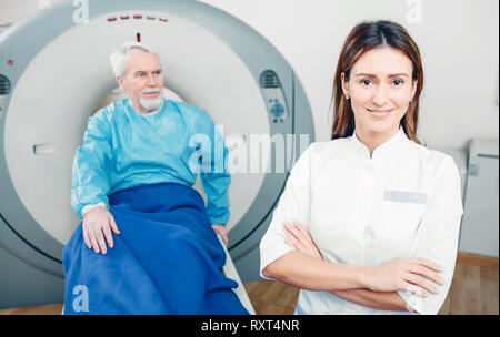 Medico radiologo, sorridere mentre guardando la fotocamera. Dietro la sua seduta del paziente senior. CT scanner in ospedale Foto Stock