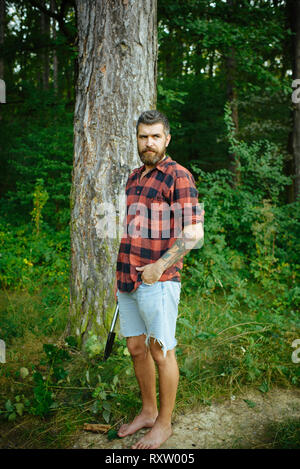 Hipster con lunga barba di verde naturale paesaggio. Uomo Barbuto con pala in foresta. Tourist in plaid shirt e Jean Corti il viaggio a piedi nudi Foto Stock