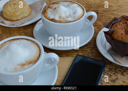 Due tazze di cappuccino su una tabella con muffin, Scone e un telefono cellulare Foto Stock