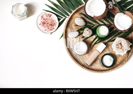 Stile composizione di bellezza. Creme per la pelle, trucco bottiglia, rose di ciottoli e pietre sul vassoio in legno Noci di cocco, palma tropicali decorazione di foglie. Prodotti cosmetici Foto Stock