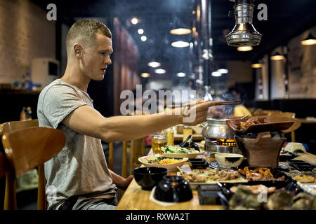 Ragazzo carino con bacchette è seduto da tavolo in legno e la cottura di pezzi di pollo su hot pot in locali del caffè tradizionale in Vietnam. Variato Foto Stock