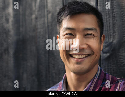 Allegro giovane uomo asiatico appoggiata contro nero parete in legno Foto Stock