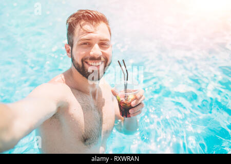 Felice l'uomo barbuto rendendo selfie in piscina. Egli sta bevendo un cocktail e rilassante. Foto Stock