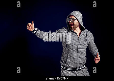 Brutale pianto barbuto ragazzo nel cofano mostra Thumbs up su sfondo nero con tendenza al blu Foto Stock