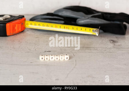 Costruzione nero guanti e un metro a nastro sullo sfondo di un muro di cemento. In primo piano l'iscrizione riparazione. Foto Stock