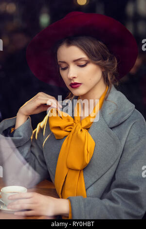 Ritratto di triste bambina indossa un floppy hat e ricoprire i. Bella bruna donna di bere il caffè nella caffetteria. Foto attraverso la finestra Foto Stock