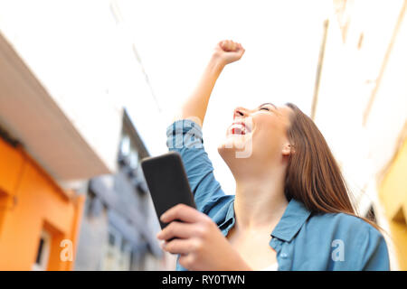 Excited donna azienda smart phone celebra il successo in strada Foto Stock