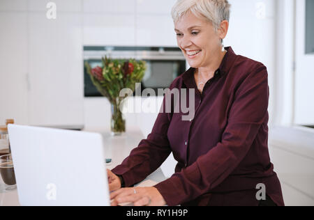 Ritratto di una bella donna matura sorridente, digitare sul proprio computer portatile, in cucina Foto Stock