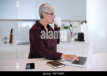 Bella donna senior utilizzando computer portatile a casa, la navigazione in internet. Signora anziana digitando su computer e gustando una tazza di caffè Foto Stock