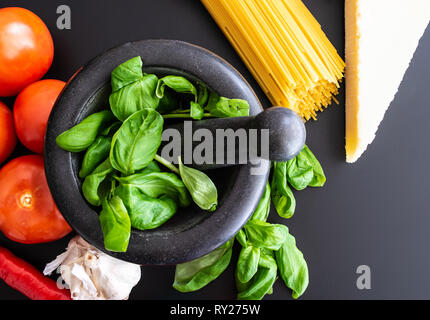 Preparare la pasta italiana con basilico fresco in un mortaio, pomodori, aglio, pepe e parmigiano on dark bancone cucina Foto Stock