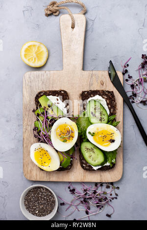Pane di segale con avocado, uovo, cetriolo, crema di formaggio e i germogli su un tagliere su sfondo grigio. vista da sopra Foto Stock