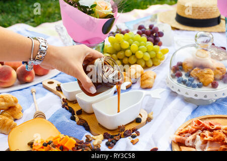 Donna mano versa il miele dalla bottiglia trasparente a sauceboat, a picnic estivo dello sfondo. Foto Stock