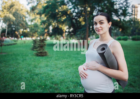 Felice giovane donna incinta stand nel parco e pongono alla fotocamera. Ella attesa yoga laminati di mate e tenere le mani sulla pancia. Modello sorriso. Foto Stock