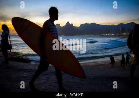 Silhouette di un surfista camminando sulla spiaggia di Ipanema boardwalk a Arpoador davanti a una vista del tramonto di due fratelli Montagna in Rio de Janeiro, Brasile Foto Stock