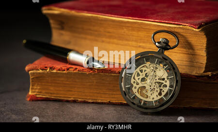 Avvolgimento pocket watch collocato accanto al vecchio libro e la penna sul libro nelle tenebre e la luce del mattino. Foto Stock