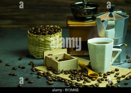 Preparare il caffè nero in tazza bianca e illuminazione di mattina. Caffè torrefatto in grani nel cestello di bambù e il cucchiaio di legno. Vintage macinino da caffè e pot. Concetto o Foto Stock