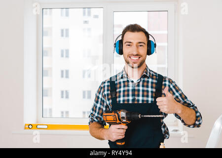 Bello tuttofare che mostra il pollice tenendo trapano in mano Foto Stock