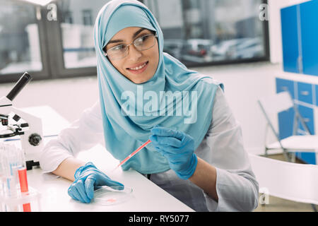 Messa a fuoco selettiva di sorridere femmina scienziato musulmano in hijab facendo prova di sangue in laboratorio Foto Stock