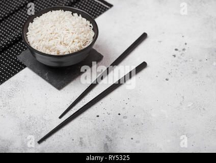 Tazza nera con bolliti basmati organico di riso al gelsomino con bacchette nere e il dolce di salsa di soia sul tappeto di pietra luce sul tavolo della cucina dello sfondo. Foto Stock