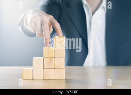 Business man walking fino passo avanti con le dita sulla carriera scala fatta da blocchi di legno; carriera o Realizzazione concetto, nei toni del blu con ligth flare Foto Stock