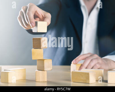 Uomo d'affari mette la prossima pietra sulla torre traballante struttura costituita da blocchi di legno; carriera o di raggiungimento o di gestione dei progetti complessi concetto, nei toni del blu Foto Stock