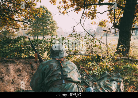 Ri-enactor vestito come il tedesco della Wehrmacht Soldato di fanteria nella guerra mondiale II seduta nascosti con un fucile arma in un agguato in trincea nella foresta di autunno Foto Stock