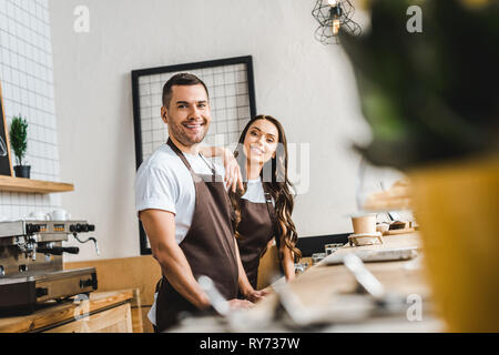 Messa a fuoco selettiva di cassieri in marrone grembiuli in piedi e sorridente dietro il bar in legno contatore in coffee house Foto Stock