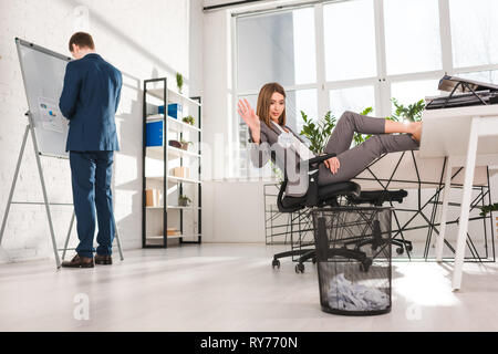 Messa a fuoco selettiva di imprenditrice seduti su una sedia e gettare la carta nel vassoio vicino a un collega, la procrastinazione concept Foto Stock