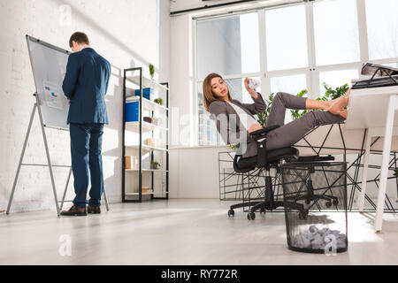 Messa a fuoco selettiva di attraenti imprenditrice seduti su una sedia e gettare la carta nel vassoio vicino a un collega, la procrastinazione concept Foto Stock