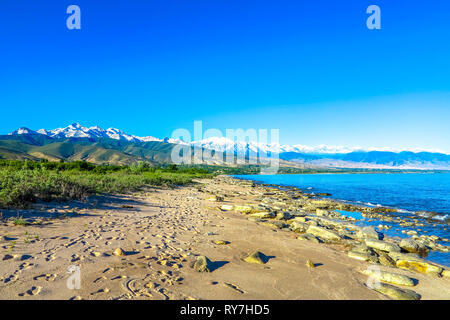 Issyk Kul Lake Shore rocce di pietra spiaggia sabbiosa con Snow capped Tian Shan Mountain Range Foto Stock